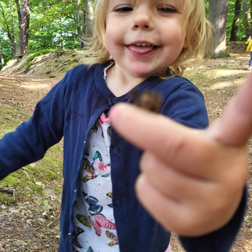 Kind lächelt im Wald und zeigt auf eine kleine Kreatur; kurze blonde Haare, blaue Jacke.