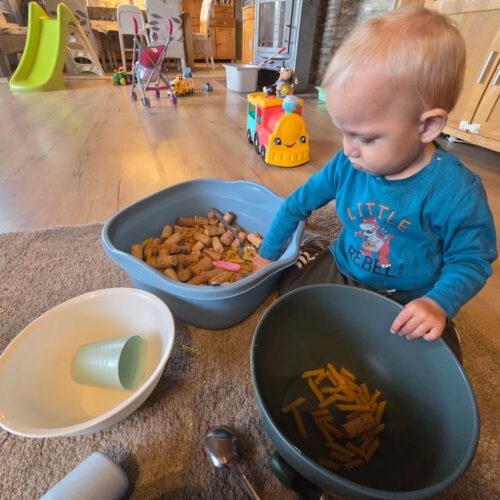 Lachendes Kleinkind in blauem Shirt spielt mit Nudeln in Schuesseln